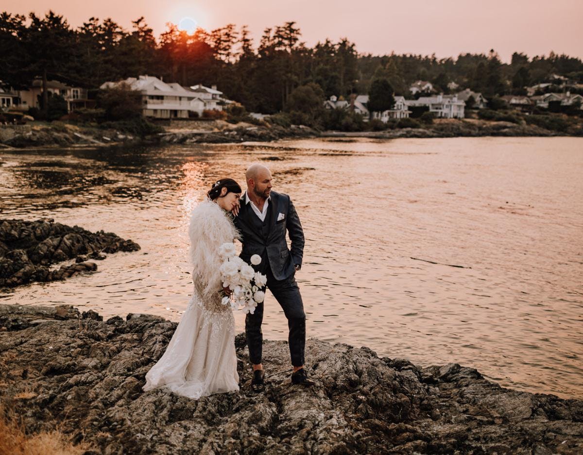 Groom and Bride with white monochrome bouquet in hand, standing on rocks alongside the ocean